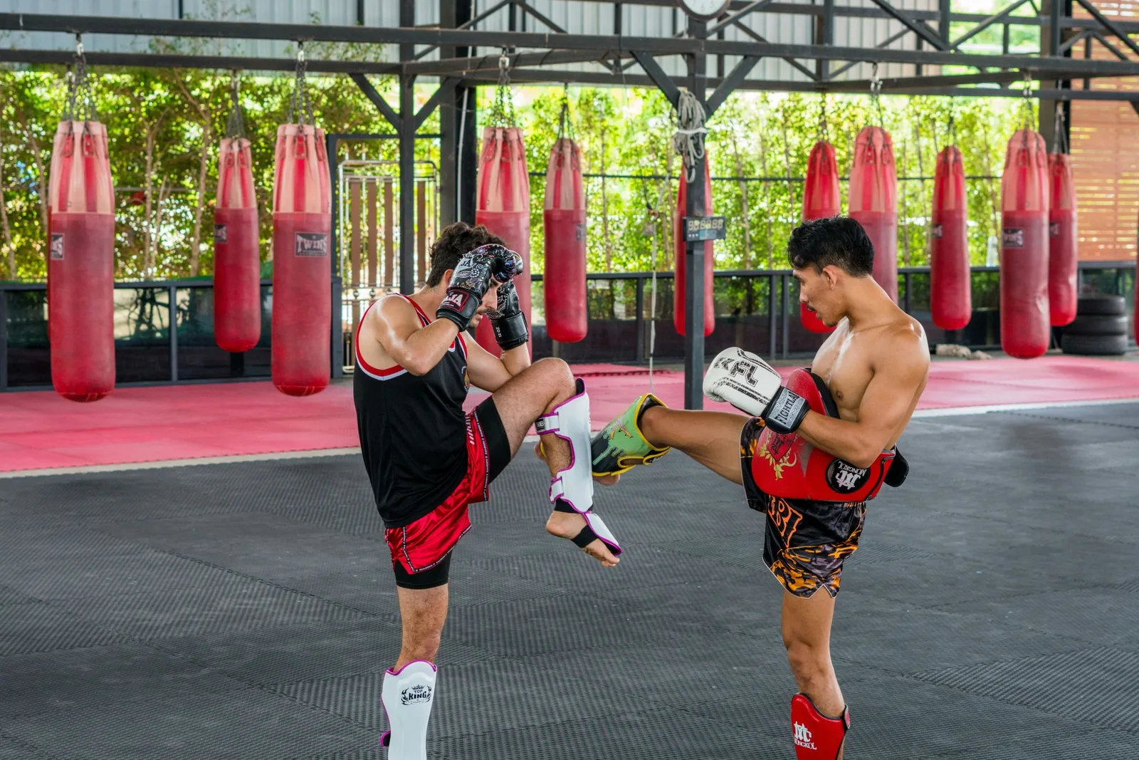 Two men practicing Muay Thai sparring in a gym with red punching bags in the background.