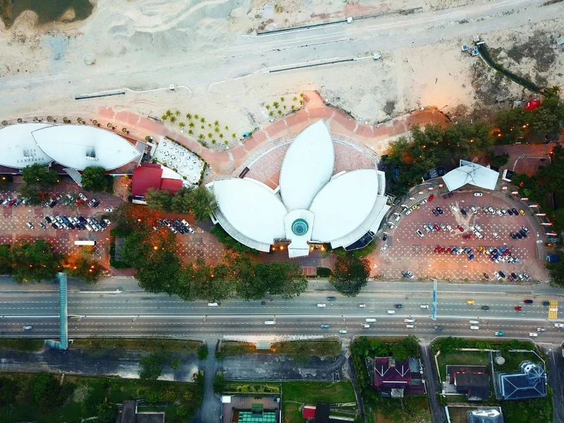 Aerial view of a lotus-shaped building in Johor Bahru, Malaysia, surrounded by parking lots and sandy construction areas near the Singapore border.