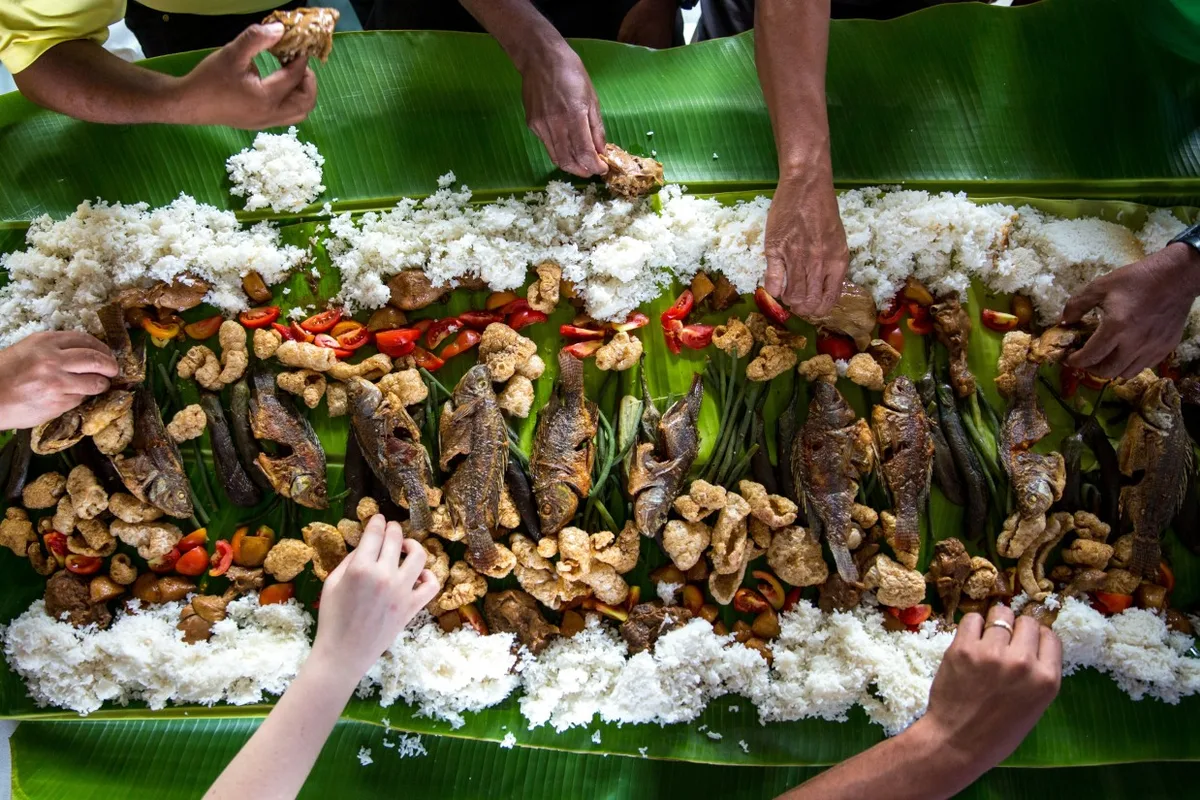 A local Thai family sharing a traditional meal of grilled fish, sticky rice, and vegetables on banana leaves in Isan.